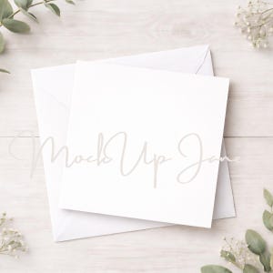 May include: A white square card and envelope, with the text "Mock Up Jane" in a light gray script, are arranged on a light wood surface. Greenery and small white flowers are placed around the card and envelope.