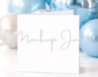 May include: A white square card with the text "Mockup Jane" in a light gray script. The card is propped up, with a few other cards behind it. In the background are several blue and silver balloons.