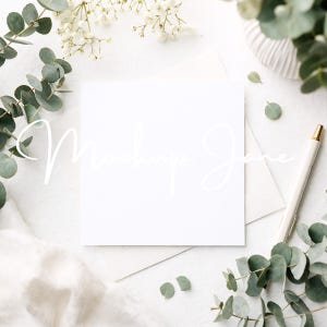 May include: A flat lay featuring a blank white square card with the text "Mockup Jane" in a delicate script. The card is surrounded by eucalyptus leaves, white flowers, a white pen, and a white envelope, all arranged on a white surface.
