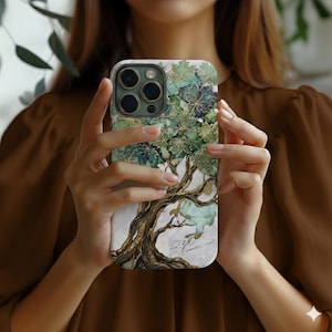 May include: A smartphone case featuring a tree design with green leaves and gold accents. The tree trunk and branches are a golden brown color, set against a white and marble-like background. The case is held in a person's hands.