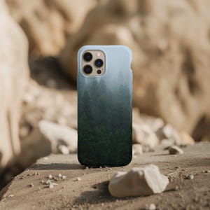 Könnte beinhalten: Hellblaue Handyhülle mit Waldmotiv. Das Design zeigt einen Farbverlauf von Hellblau nach Dunkelgrün, der einen nebligen Wald darstellt. Die Handyhülle hat einen schützenden Stoßfänger.