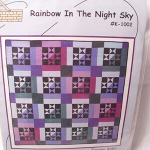 May include: A package for a quilt project titled "Rainbow In The Night Sky" by Tammy Kelly. The quilt design features a grid of squares in shades of purple, pink, and teal, with star-like patterns in the center of each square. The package is labeled "Common Threads".