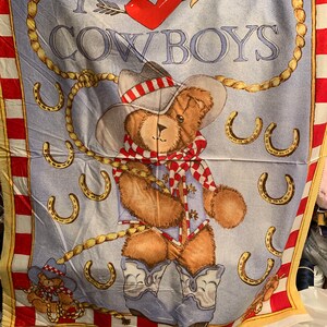 May include: A blue and yellow fabric panel with a teddy bear wearing a cowboy hat and boots. The panel says "I love cowboys" with a red heart and arrow. There are gold horseshoes around the bear.