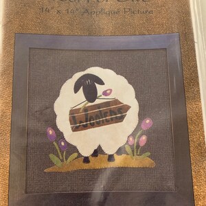May include: A white sheep with black legs and a black head carrying a wooden sign that says "Woolens". The sheep is surrounded by purple flowers and green grass. The image is framed in a black frame and is on a brown burlap background. The title "Wool For Ewe" is written above the image. The image is 14 inches by 14 inches.