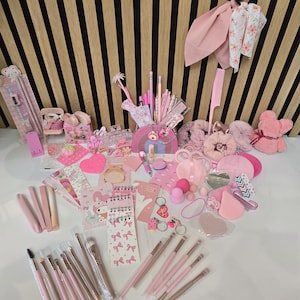 May include: A collection of pink and light pink beauty and stationery items, including makeup brushes, hair accessories, notebooks, and pencils. The items are arranged on a white surface with a striped wooden background.