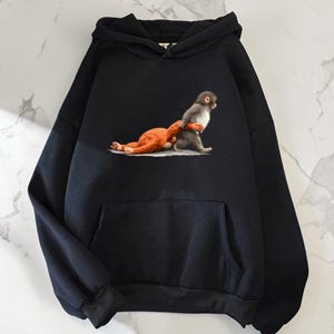 Puede incluir: Sudadera con capucha negra con bolsillo de canguro y capucha con cordón. La parte delantera presenta un gráfico de un mono gris y blanco que lleva un peluche naranja. El diseño está centrado en el pecho.