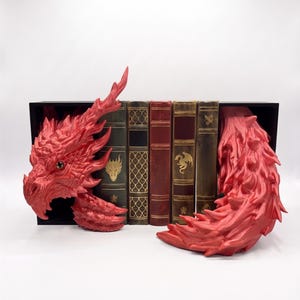 May include: A pair of red dragon bookends featuring a detailed dragon head on the left and a spiky dragon tail on the right, framing a row of four vintage-style books with gold embossed dragon and geometric patterns on their spines.