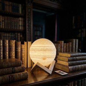 Op de afbeelding: Een gloeiende, bolvormige lamp die op Jupiter lijkt, met lichte en donkere banden, staat op een houten standaard. De lamp staat op een donkere houten plank met oude boeken in een bibliotheek. Een afstandsbediening is zichtbaar.