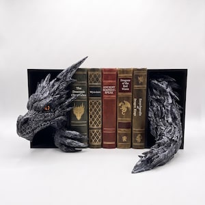 Può includere: Un set di fermalibri a tema drago nero con una testa di drago dettagliata a sinistra e una coda squamosa a destra, che incornicia cinque libri con titoli tra cui The Draconic Chronicles, Wym-lore, Ancient Serpent-Speak e Dragon Kings.