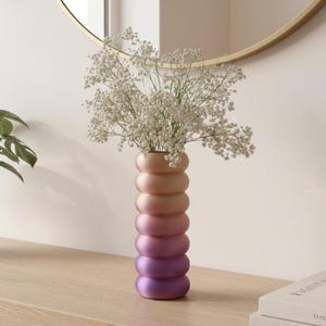 May include: A decorative vase with a gradient colour scheme, transitioning from peach to purple. The vase has a unique stacked ring design and holds a bouquet of white flowers. The vase is displayed on a wooden surface.