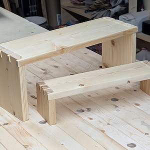 May include: Two unfinished wooden step stools. The larger stool is rectangular with a flat top and four legs. The smaller stool is also rectangular with a flat top and four legs. Both stools are made of light-colored wood and are placed on a wooden surface.