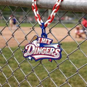 May include: A baseball-themed pendant with the text "I Hit Dingers" in a stylized font, hanging from a chain-link fence. The pendant is blue, red, and white, with a baseball bat and ball graphic. The chain is red and white.