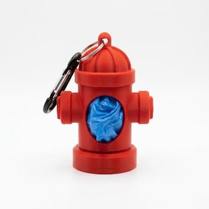 May include: A red, fire hydrant-shaped dog waste bag dispenser with a black carabiner clip. The dispenser holds a blue bag and is designed to attach to a leash or bag. The product is made of plastic.
