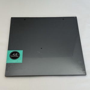 May include: A square black chalkboard surface wrapped in clear plastic. A teal rectangular label in the bottom left corner features the white text Chalk Couture and Couture Boutique 9" x 9" Board. Three small circular holes are visible at the top.