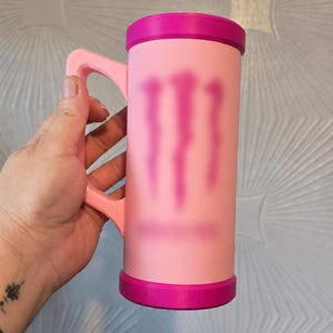 May include: A cylindrical mug with a light pink body and a bright magenta rim and base. The mug features a matching light pink handle and a blurred magenta graphic design on the front surface, resembling a stylised claw mark logo.