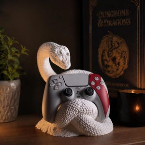 May include: A white, coiled snake-shaped controller stand holding a grey and red video game controller. The snake has a textured, scale-like surface and is positioned in front of a black Dungeons & Dragons book with gold lettering.