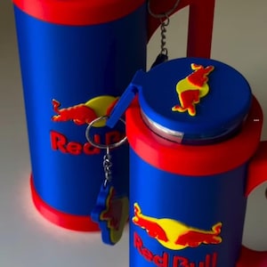 May include: Two blue and red cylindrical beverage can holders with handles. Each holder features the Red Bull logo in yellow and red, with a matching emblem on the lid. A small keychain with a Red Bull logo is attached to the handle of the smaller holder.