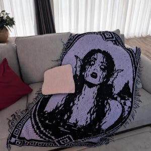 May include: A rectangular crochet throw blanket featuring a black and light purple graphic design of a figure with long hair and pointed ears. The blanket has a decorative border with geometric patterns and fringe along the edges.