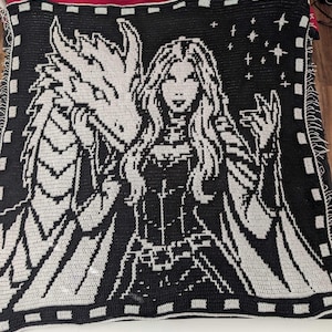 May include: A rectangular crochet blanket featuring a high-contrast black and white graphic design. The pattern depicts a long-haired figure standing beside a large dragon, with a cluster of stars in the upper right corner and a border of squares.