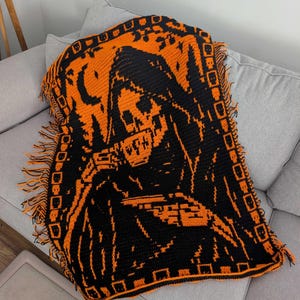 Op de afbeelding: Een rechthoekige gehaakte deken met een contrastrijk zwart en oranje ontwerp van een figuur met een kap die uit een kopje drinkt. De rand is versierd met een herhalend schedelpatroon en de zijkanten zijn afgewerkt met korte franjes.