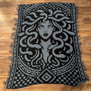 May include: A black and blue crocheted blanket featuring a Medusa design. The blanket has a detailed pattern of Medusa's face and snakes, with a geometric border and fringe. The color scheme is primarily black and blue.