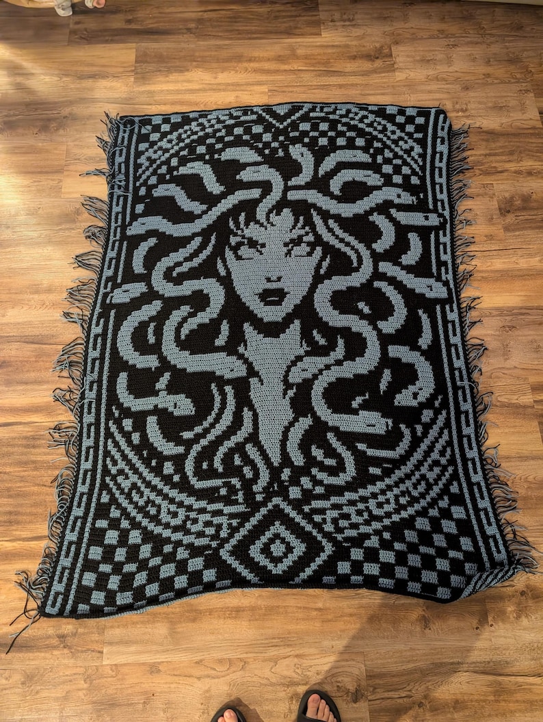 May include: A black and blue crocheted blanket featuring a Medusa design. The blanket has a detailed pattern of Medusa's face and snakes, with a geometric border and fringe. The color scheme is primarily black and blue.