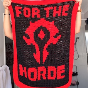May include: A handmade crochet blanket in black and red, featuring the words "FOR THE HORDE" and a stylized Horde symbol. The blanket is held up, showcasing the bold design and color scheme.