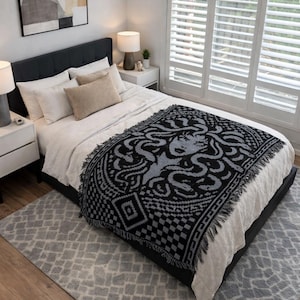 May include: A black and gray woven throw blanket with a Medusa design is draped over a bed with white bedding. The blanket has a fringed edge and is placed on a gray rug. The room has white walls and a large window.