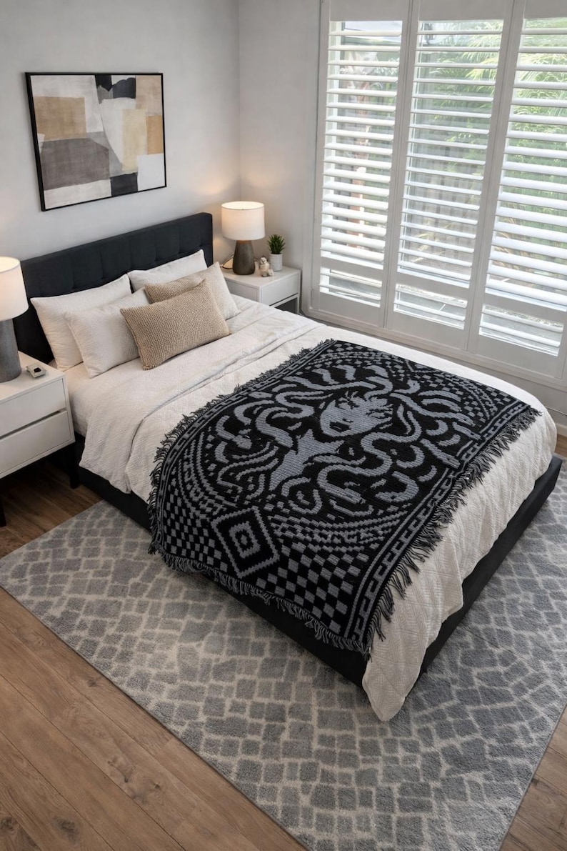 May include: A black and gray woven throw blanket with a Medusa design is draped over a bed with white bedding. The blanket has a fringed edge and is placed on a gray rug. The room has white walls and a large window.