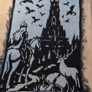 May include: A rectangular crochet blanket featuring a black and light blue design of a tall castle, a figure on horseback, a deer, and several flying dragons. The edges of the blanket have a fringed border with alternating black and light blue yarn.