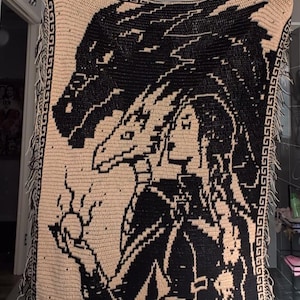 Può includere: Una coperta rettangolare all'uncinetto con un design bicolore nero e beige. Il motivo raffigura un grande drago che sovrasta una persona che tiene in mano un globo luminoso. La coperta è rifinita con un bordo a greca geometrica e frange.