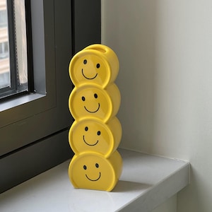 Puede incluir: Un jarrón de cerámica amarilla con un diseño de caras sonrientes apiladas. El jarrón está compuesto por cuatro secciones redondeadas, cada una con una cara sonriente negra. El jarrón se exhibe en un alféizar blanco cerca de un marco de ventana oscuro.