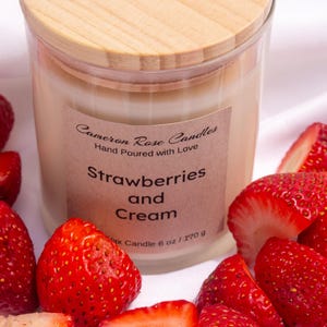 Puede incluir: Una vela en frasco de vidrio transparente con cera clara, tapa de madera y etiqueta color canela que dice Cameron Rose Candles, Hand Poured with Love, Strawberries and Cream, Wax Candle 6 oz / 170 g, rodeada de numerosas fresas rojas frescas.