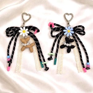 May include: Two decorative keychains with black and white rope bows, daisy and flower charms, and heart-shaped clasps. One keychain has a brown balloon dog charm, the other a black balloon flower. Both have lace and colorful beads.