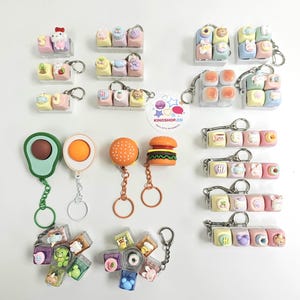 May include: Collection of colorful keychains featuring various designs, including food items like a burger and avocado, and cute characters. Some keychains have the text "KINOSHOP.CO".
