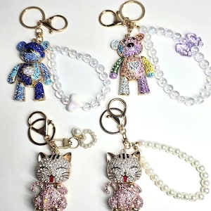 May include: Four keychains featuring two teddy bears and two cats covered in colorful rhinestones. The teddy bears are blue and multicolored, while the cats are pink and silver. Each keychain includes a gold metal ring and a beaded wrist strap.