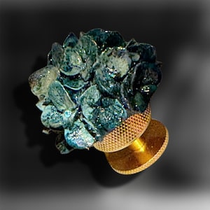 May include: A decorative cabinet knob featuring a cluster of dark teal and blue hydrangea-like petals with a subtle shimmer. The base of the knob is made of polished gold-toned metal with a textured, cross-hatched pattern on the circular mounting plate.