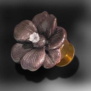 Puede incluir: Un pomo con forma de flor de cinco pétalos en color morado oscuro con acabado brillante y un pequeño detalle central en blanco translúcido. La flor está unida a una base metálica cilíndrica y texturizada de color dorado en la parte posterior.