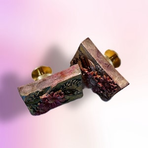 May include: Two decorative, rectangular drawer pulls with floral designs. The pulls are a mix of brown, gold, and burgundy, with a textured appearance. Each pull has a gold-coloured knob for easy handling.