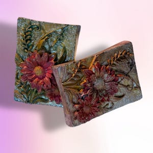 Puede incluir: Dos piezas decorativas rectangulares texturizadas con un diseño floral. Las piezas presentan flores rojo-burdeos en relieve y follaje verde y dorado. El fondo tiene un tono moteado y terroso, y el efecto general es rústico y artístico.