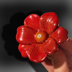 Op de afbeelding: Een glanzende, felrode bloemvormige broche met vijf afgeronde bloemblaadjes met fijne, stralende textuurlijnen. Het midden van de bloem is een gladde, goudgele ronde schijf met een enkele, kleine, heldere, sprankelende edelsteen in het midden.