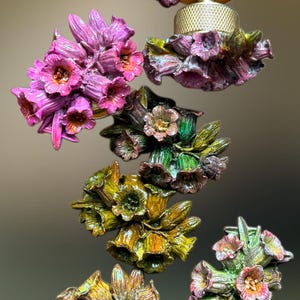 May include: A collection of six decorative cabinet knobs shaped like clusters of bell-shaped flowers in various colours including deep purple, bright pink, green, and yellow, each featuring a gold-toned metal base with a textured cylindrical knob.