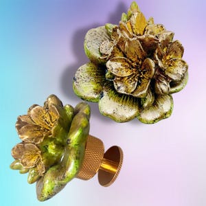 Puede incluir: Dos pomos decorativos para armarios con forma de flores en flor. Cada pomo presenta pétalos en capas con un acabado dorado metálico, acentuado por motas verdes y negras. Una base cilíndrica texturizada se conecta a una placa trasera.