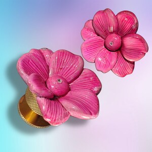 Op de afbeelding: Twee oorplugs met felroze, vijfbladige bloemdesigns met textuur. Elke bloem heeft een kleine, heldere steen in het midden. De plugs hebben een goudkleurige, cilindrische basis met een gekartelde, kruislings gearceerde rand voor grip.