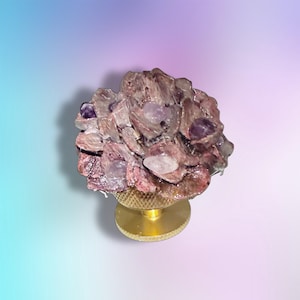 May include: A decorative object featuring a cluster of translucent, irregular purple and clear crystal-like shards arranged in a floral shape, mounted atop a circular, textured gold-toned metal base with a flat, smooth pedestal stand.