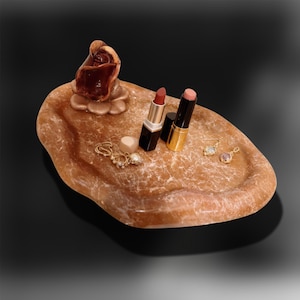 Könnte beinhalten: Eine braune Steinschale mit einer geschnitzten Rose und verschiedenen Accessoires. Die Schale enthält zwei Lippenstifte, ein Paar Ohrringe, ein kleines herzförmiges Objekt und eine goldene Halskette mit einer Perle. Die Schale steht auf einer schwarzen Oberfläche.