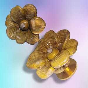 May include: Two flower-shaped cabinet knobs with a mustard yellow, glazed finish and textured petals. Each knob features a central, small, rough, silver-toned metallic accent. The base of the lower knob is a circular, gold-toned metal hardware piece.