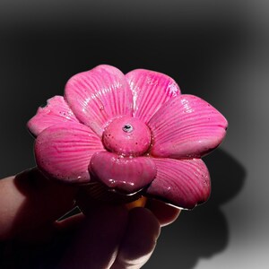 May include: A bright pink flower-shaped knob with a glossy, textured finish and a small central indentation. The petals feature subtle radiating lines and a slightly uneven, organic surface texture, creating a distinct three-dimensional appearance.
