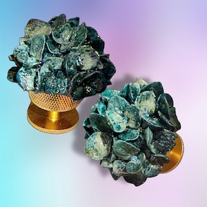 May include: Two decorative knobs featuring sculpted dark teal and blue hydrangea flower heads with small embedded crystals. Each floral cluster is mounted on a circular, textured gold-toned metal base with a knurled edge for a secure grip.