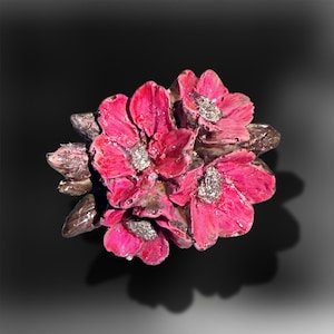 May include: A decorative brooch featuring a cluster of three vibrant pink flowers with dark brown, glossy leaves and textured, metallic silver centres. The floral arrangement is crafted with a high-gloss finish and detailed, layered petals.